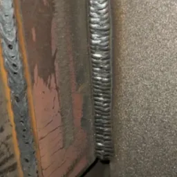 Precyzyjne spawy łączące metalowe elementy pod kątem prostym, ukazujące regularny wzór spoiny. Widoczne ślady obróbki i chropowata faktura metalu.