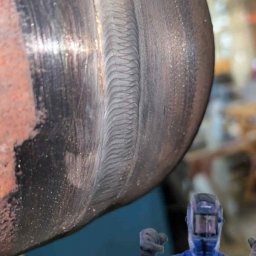 Klaudiusz Brzóska WELD - Precyzyjne spawy na metalowej rurze, widoczny wzór spoiny. W tle postać w kombinezonie i masce ochronnej.