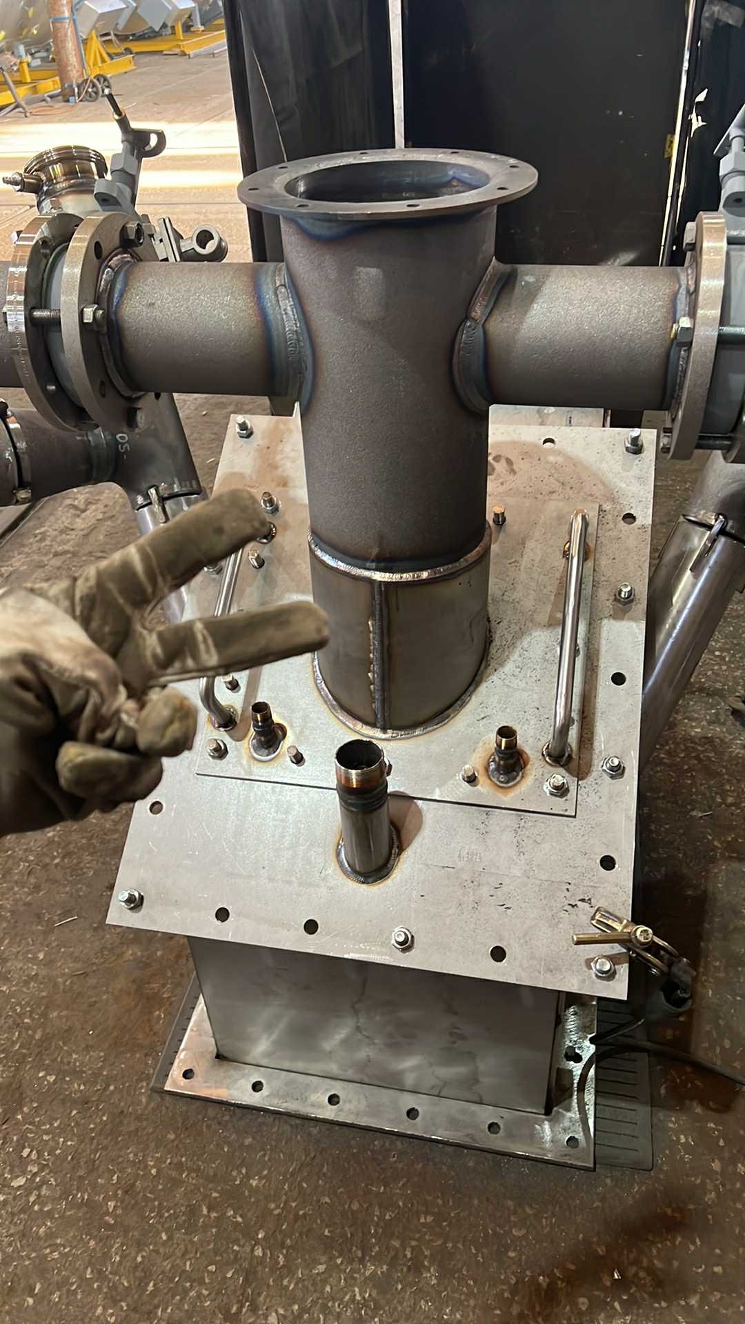 Metalowa konstrukcja z rur i blach, spawana, z widocznymi śladami obróbki. Dłoń w rękawicy pokazuje znak zwycięstwa. Elementy połączone śrubami.