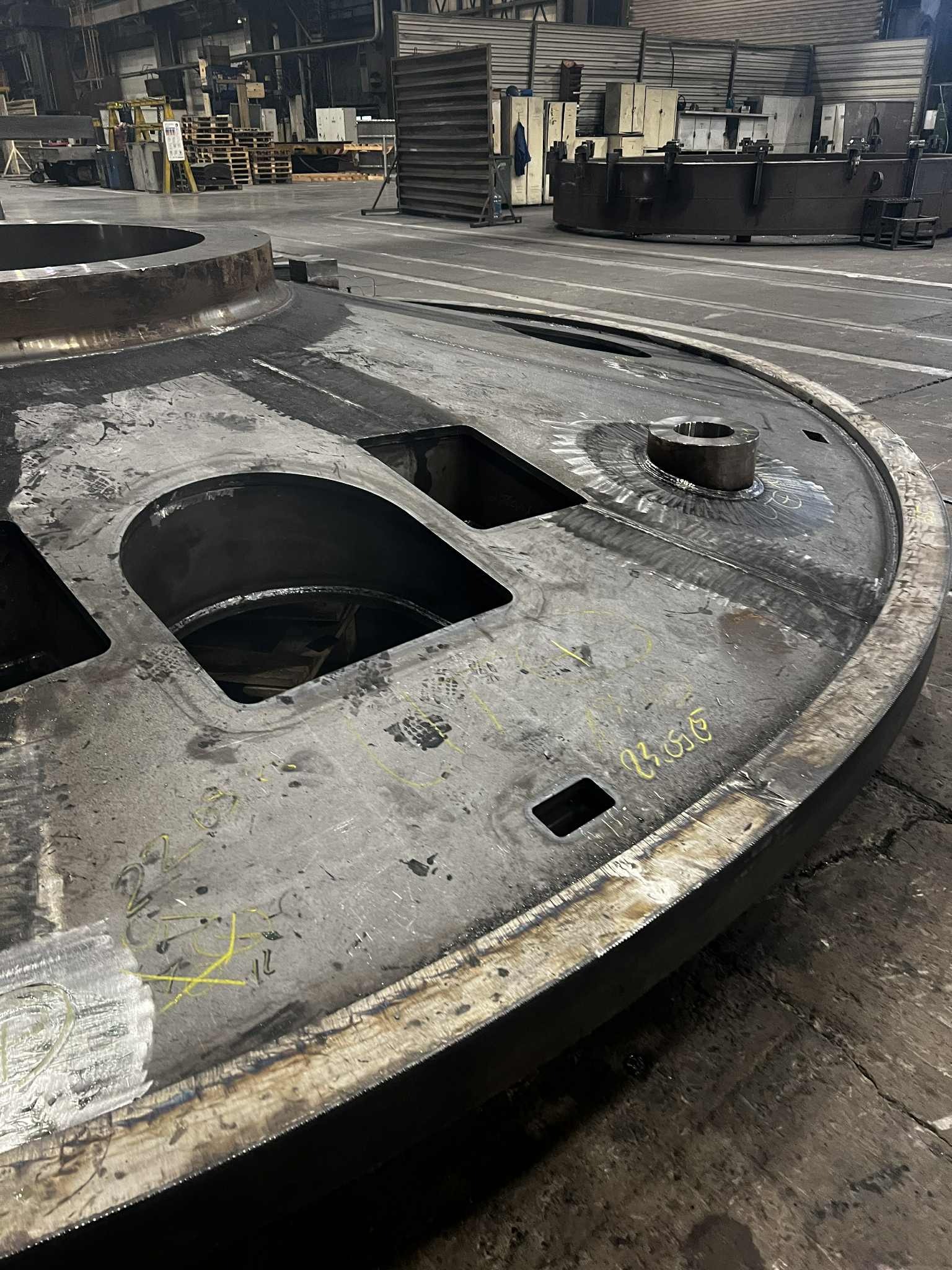 Obrabiany, metalowy element o okrągłym kształcie z otworami, leżący na podłodze hali produkcyjnej. Widoczne ślady obróbki, spawania oraz żółte oznaczenia na powierzchni.