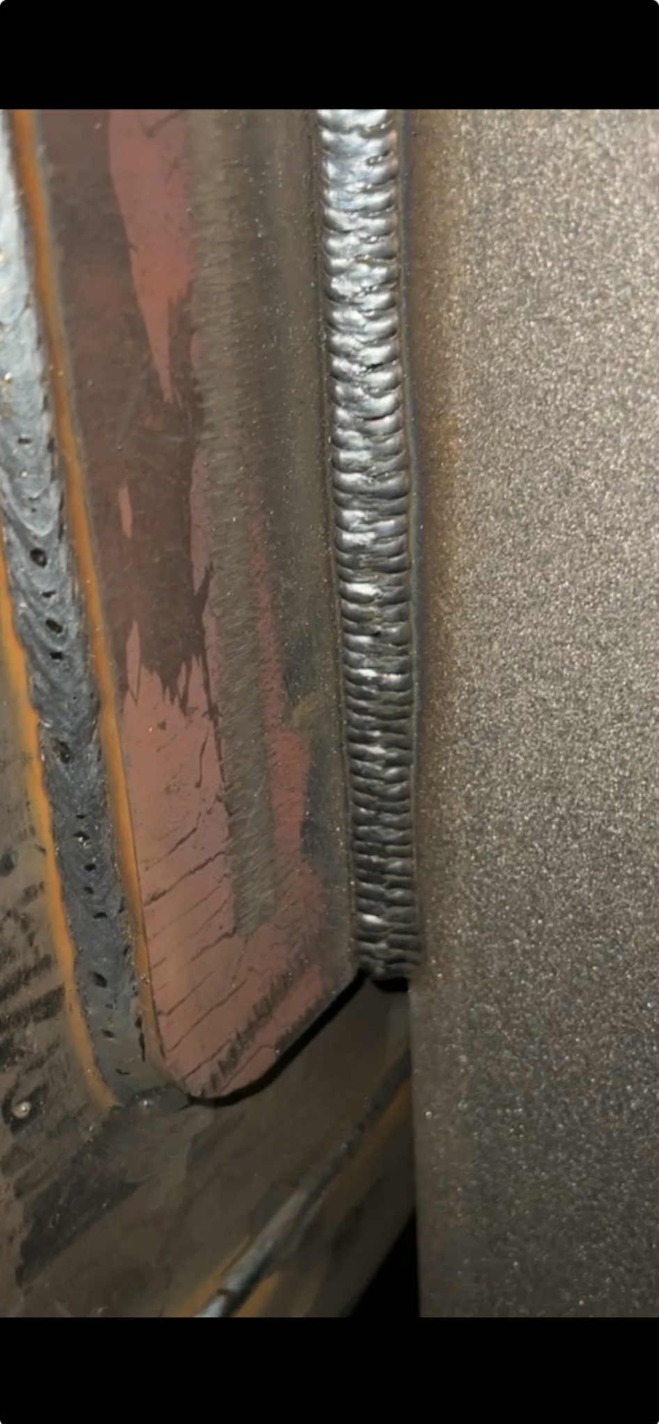 Precyzyjne spawy łączące metalowe elementy pod kątem prostym, ukazujące regularny wzór spoiny. Widoczne ślady obróbki i chropowata faktura metalu.