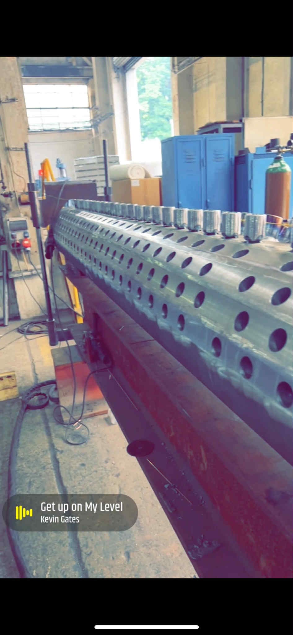 Metalowy cylinder z otworami i wspawanymi tulejami, umieszczony na czerwonej konstrukcji w hali produkcyjnej. Widoczne narzędzia i butla z gazem.