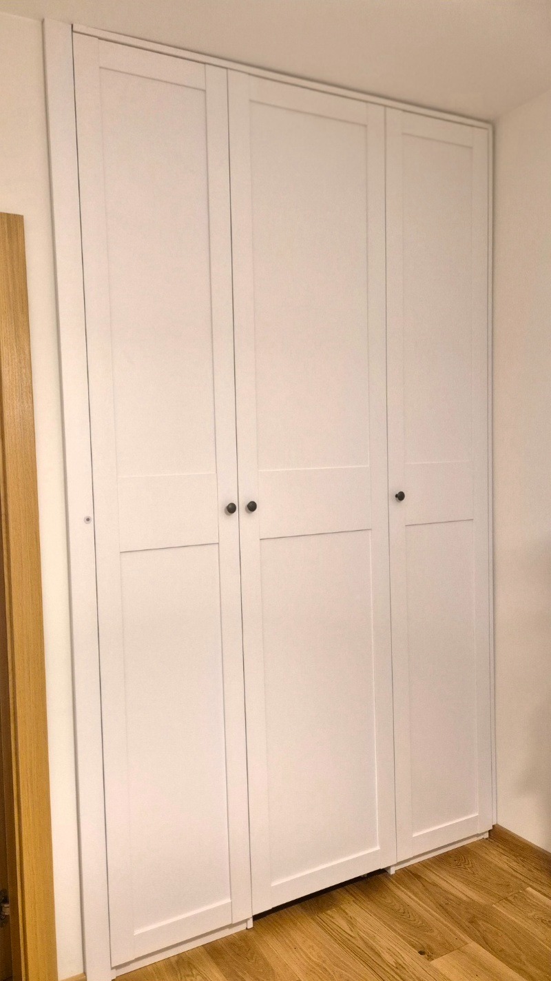 Biała szafa wnękowa z trzema parami drzwi, widoczna na tle jasnej ściany i drewnianej podłogi. Minimalistyczny design, ciemne okrągłe uchwyty.