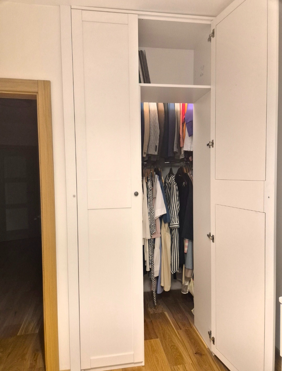 Biała szafa wnękowa z otwartymi drzwiami, ukazująca ubrania wiszące w środku. Widoczna drewniana podłoga i fragment ościeżnicy drzwiowej.