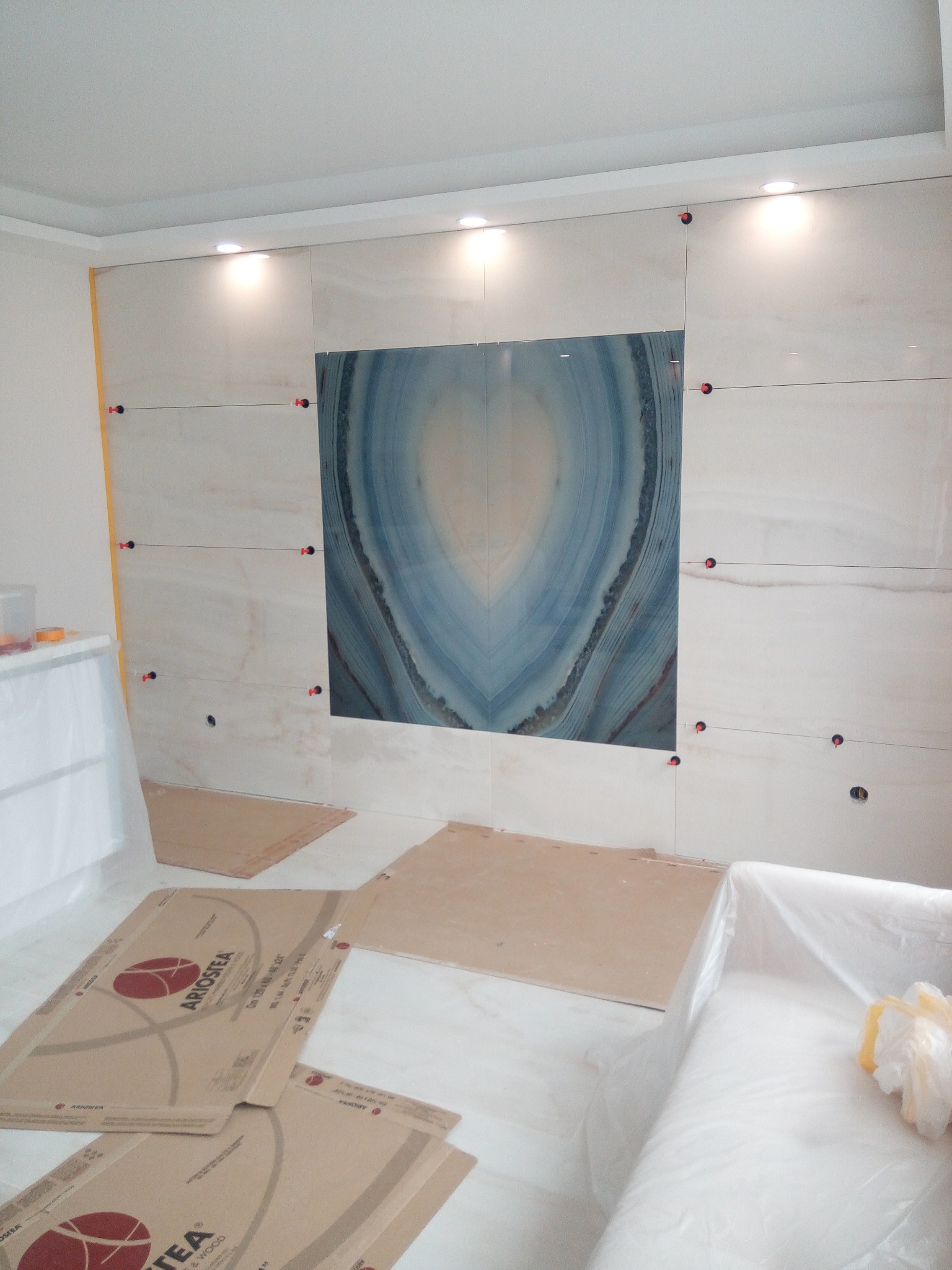 Montaż płytek imitujących agat na ścianie w łazience, z użyciem poziomicy i krzyżyków dystansowych. Podłoga zabezpieczona kartonami.