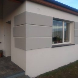 FIRMA BUDOWLANA MATEUSZ GRONOWICZ - Wykończona elewacja domu jednorodzinnego z dekoracyjnymi, szarymi elementami w formie prostokątów wokół okna i na rogu budynku. Widoczny fragment wejścia i okna.