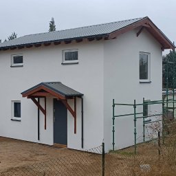 FIRMA BUDOWLANA MATEUSZ GRONOWICZ - Otynkowany dom z szarą stolarką i zadaszeniem nad wejściem. Na elewacji widoczne rusztowanie. Budynek otoczony zielenią i niskim ogrodzeniem.