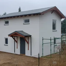 Otynkowany dom z szarą stolarką i zadaszeniem nad wejściem. Na elewacji widoczne rusztowanie. Budynek otoczony zielenią i niskim ogrodzeniem.