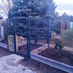 FIRMA BUDOWLANA MATEUSZ GRONOWICZ - Nowoczesne ogrodzenie panelowe z podmurówką betonową i furtką, widok z zewnątrz posesji, na tle zieleni i zabudowań gospodarczych. Szara kolorystyka.