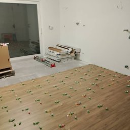 FIRMA BUDOWLANA MATEUSZ GRONOWICZ - Układanie płytek podłogowych w Glinno, z użyciem systemu poziomowania. W tle widoczne paczki płytek i narzędzia.