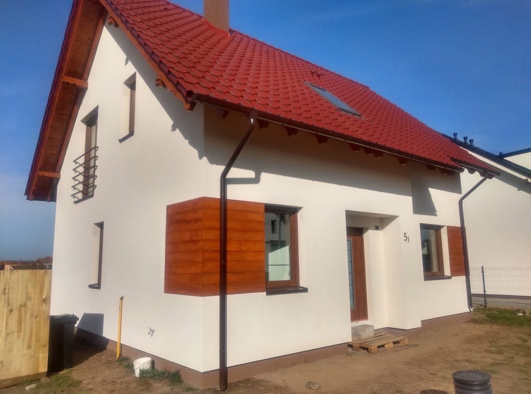 Dom jednorodzinny z czerwoną dachówką i jasną elewacją. Okna z drewnianymi okiennicami i numerem 51 nad drzwiami. Widoczne rynny i fragment ogrodu.