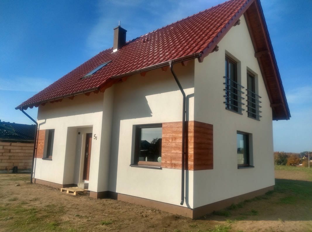 Nowoczesny dom jednorodzinny po renowacji elewacji, z brązową stolarką okienną i dachem z czerwonej dachówki. Widoczne detale wykończenia.