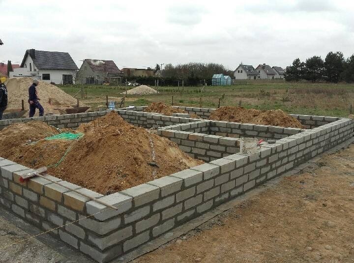 Wznoszenie fundamentów z bloczków betonowych, widoczne hałdy ziemi wewnątrz. W tle domy jednorodzinne i pochmurne niebo.