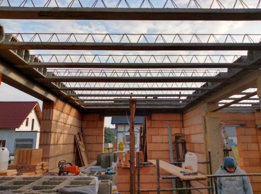 Wnętrze surowego budynku w trakcie budowy. Widoczne ceglane ściany, stalowe belki stropowe i narzędzia budowlane. W tle dom z czerwonym dachem.