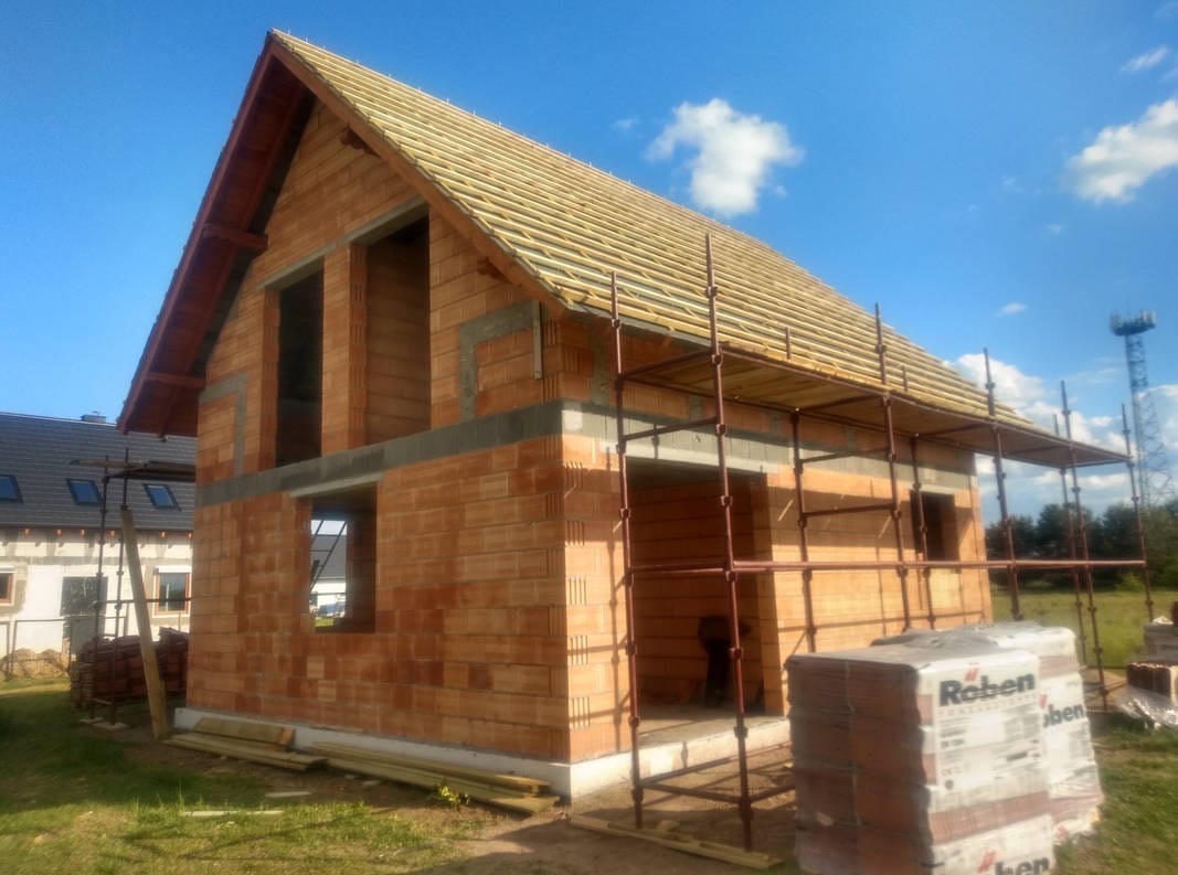 Surowy, dwukondygnacyjny dom z czerwonej cegły w trakcie budowy, z widocznym rusztowaniem i elementami konstrukcji dachu na tle błękitnego nieba.