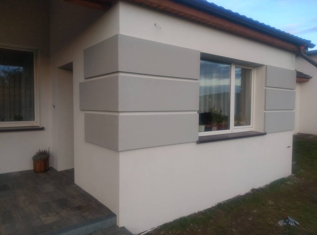 Wykończona elewacja domu jednorodzinnego z dekoracyjnymi, szarymi elementami w formie prostokątów wokół okna i na rogu budynku. Widoczny fragment wejścia i okna.