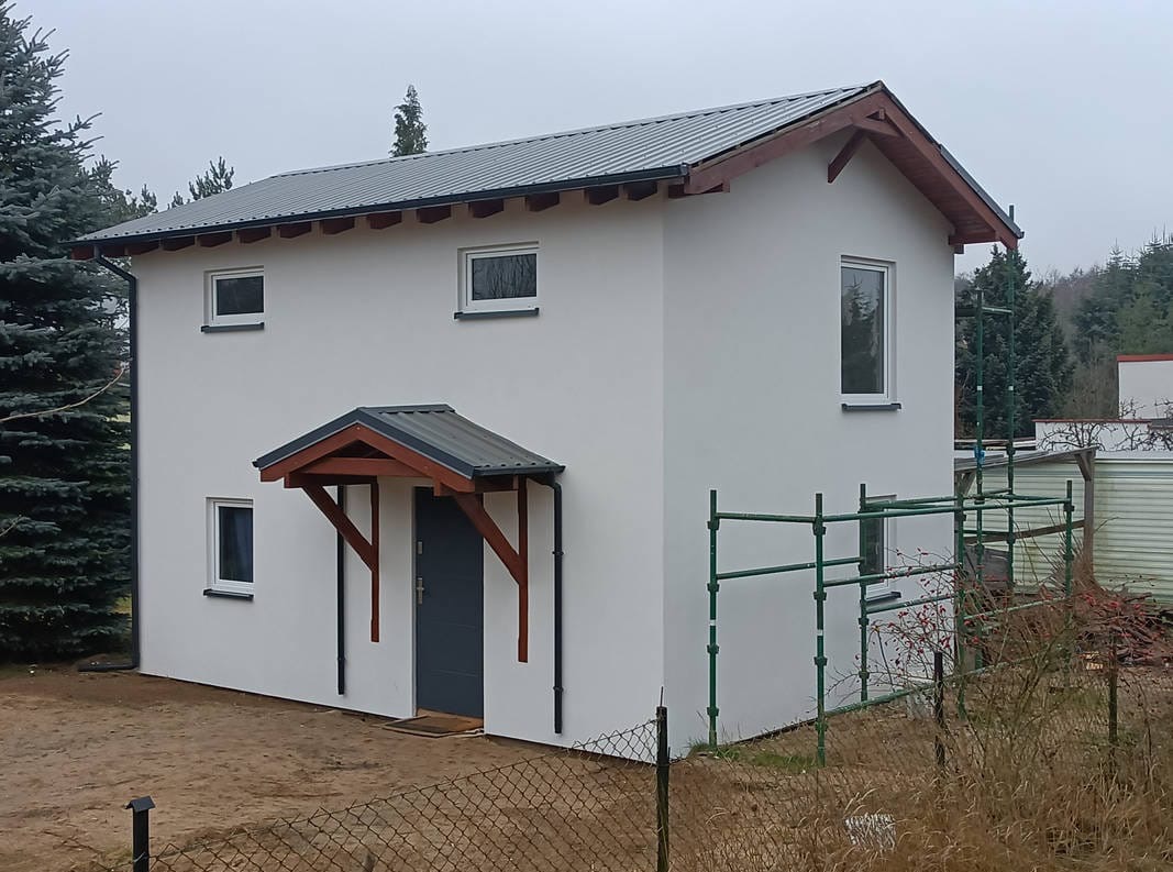 Otynkowany dom z szarą stolarką i zadaszeniem nad wejściem. Na elewacji widoczne rusztowanie. Budynek otoczony zielenią i niskim ogrodzeniem.