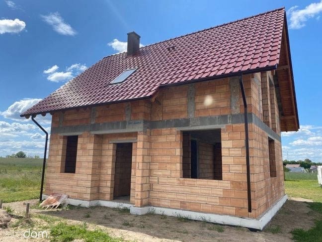 Dwukondygnacyjny dom w trakcie budowy z czerwonej cegły, z dachem pokrytym czerwoną dachówką, na tle błękitnego nieba z chmurami i zieleni. Widoczne niezadaszone okna.