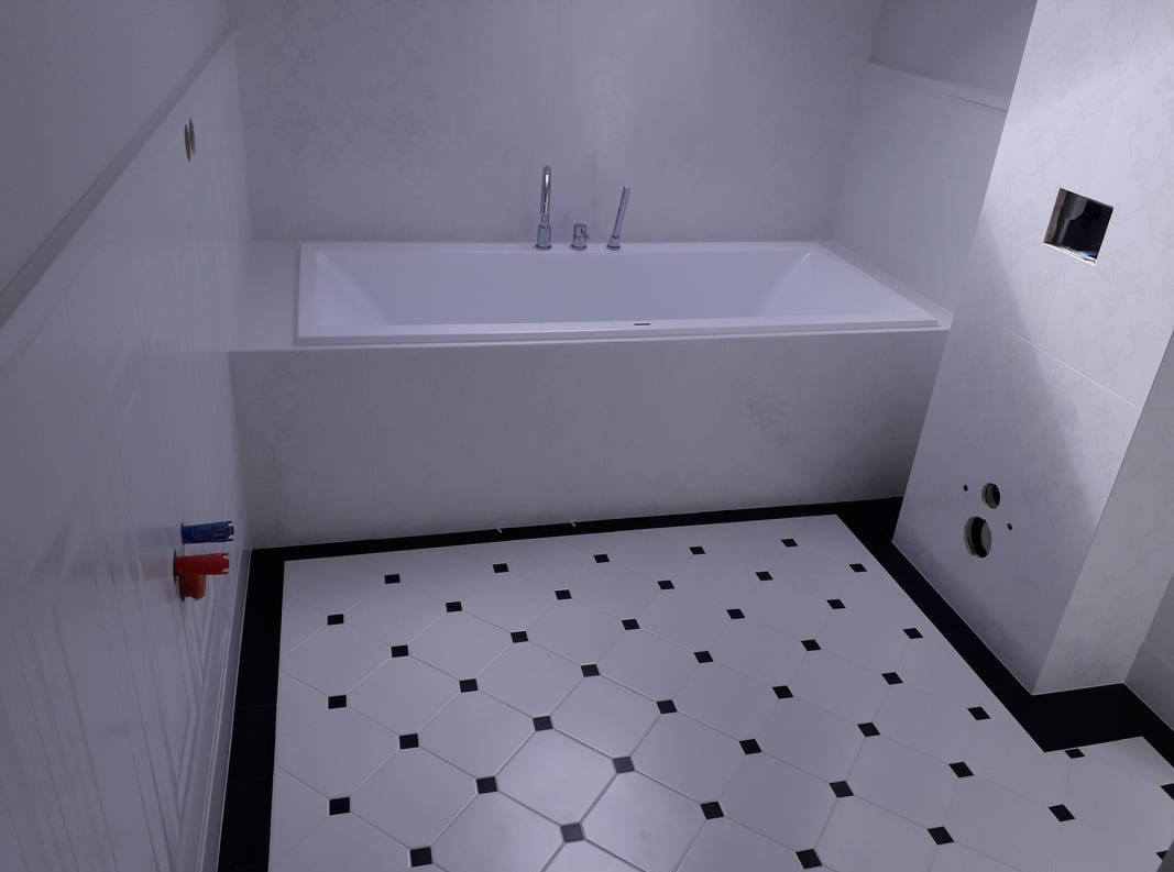Minimalistyczna łazienka z białą wanną i wzorzystą podłogą w romby z czarnymi akcentami. Widoczne przyłącza wodne i fragment instalacji.