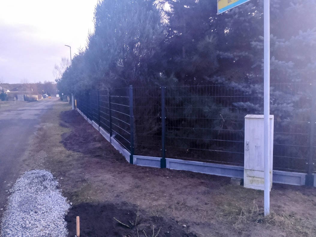 Nowoczesne, metalowe ogrodzenie panelowe z podmurówką betonową, oddzielające posesję od drogi. W tle drzewa iglaste i słup z tabliczką uliczną.