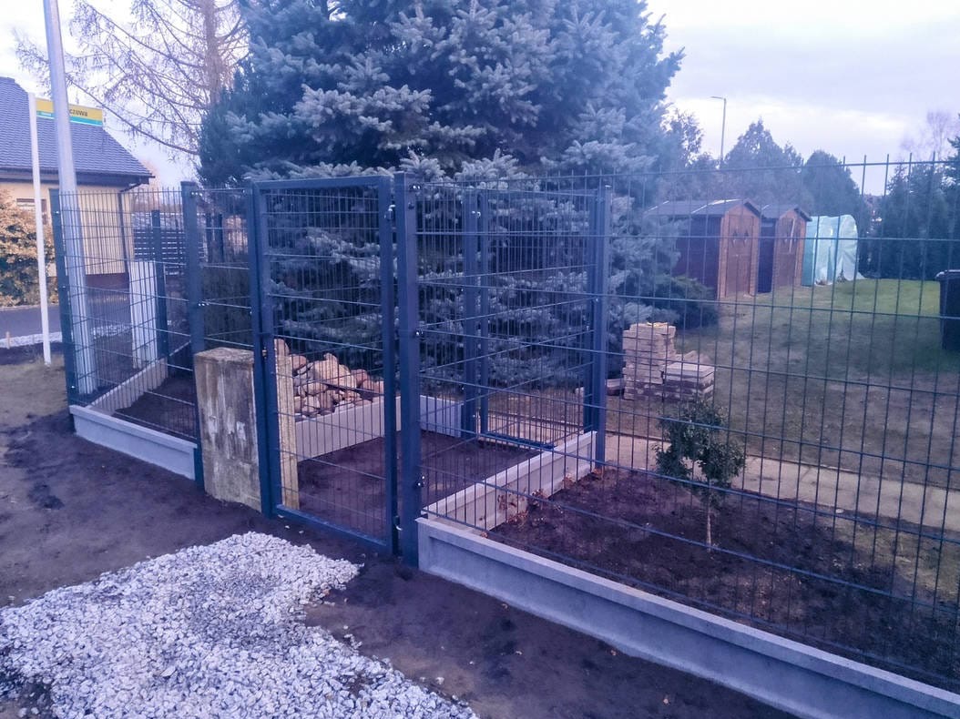 Nowoczesne ogrodzenie panelowe z podmurówką betonową i furtką, widok z zewnątrz posesji, na tle zieleni i zabudowań gospodarczych. Szara kolorystyka.