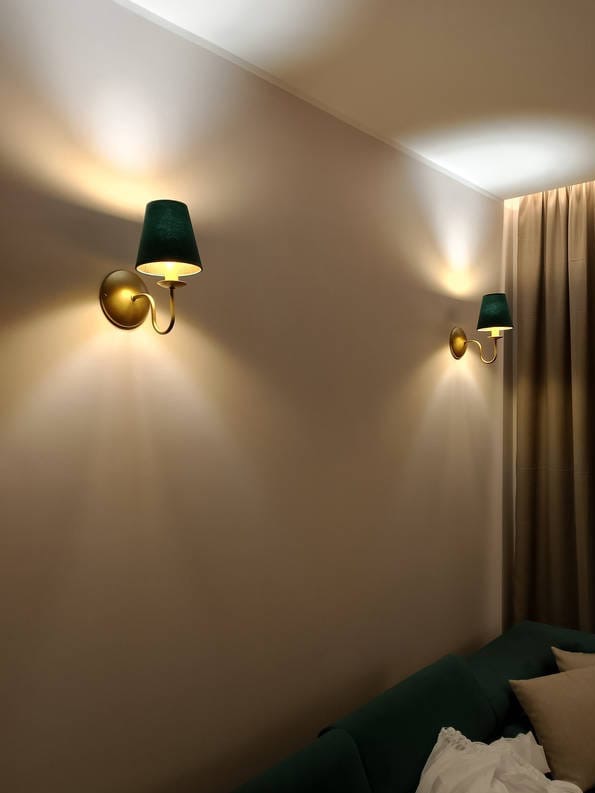 Dwa kinkiety z zielonymi abażurami i złotymi podstawami oświetlają beżową ścianę w pokoju. Widoczny fragment sofy i zasłony.
