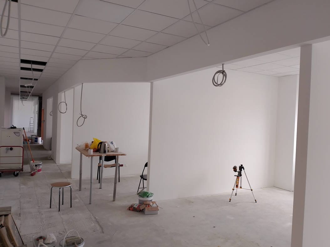 Wnętrze biura w trakcie malowania ścian na biało. Widoczne narzędzia, sufit kasetonowy, niezabezpieczone kable, stół z czajnikiem i poziomica laserowa na statywie.