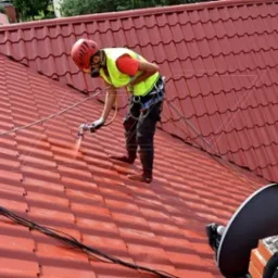 Pracownik w kasku i masce ochronnej, zabezpieczony linami, maluje czerwoną blachodachówkę na skośnym dachu za pomocą pistoletu natryskowego; widoczny komin i antena satelitarna.