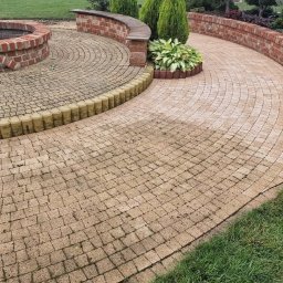 Kam-Dach Kamil Parczewski - Ogród z kostki brukowej w odcieniach beżu, z okrągłym paleniskiem z cegły i niskim murem. Widoczne elementy zieleni i oświetlenie ogrodowe.