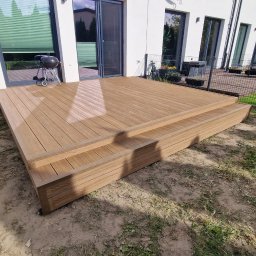 Kam-Dach Kamil Parczewski - Nowoczesny taras kompozytowy z dwoma stopniami, wykonany z desek w odcieniach brązu, na tle białego budynku z dużymi oknami i zielonego trawnika.