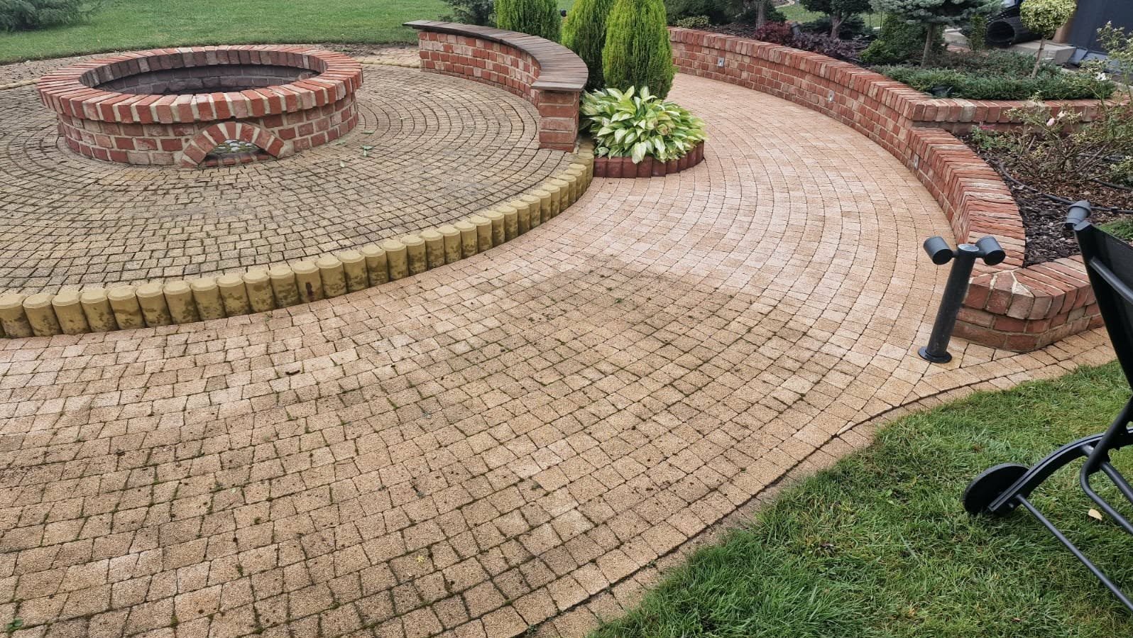 Ogród z kostki brukowej w odcieniach beżu, z okrągłym paleniskiem z cegły i niskim murem. Widoczne elementy zieleni i oświetlenie ogrodowe.