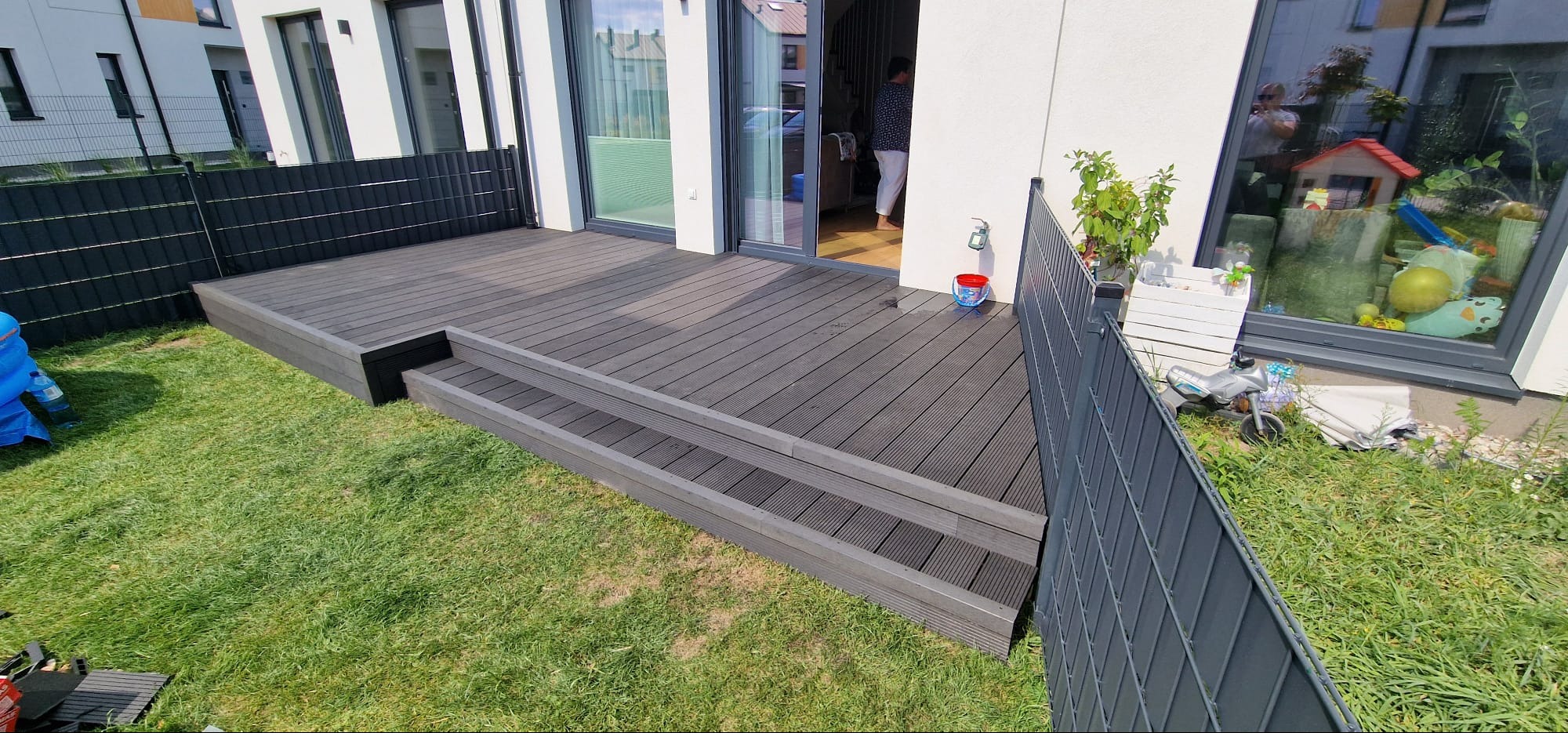 Nowoczesny taras kompozytowy z dwustopniowymi schodami, przylegający do domu z dużym oknem i przesuwnymi drzwiami. Widoczne ogrodzenie i trawnik.