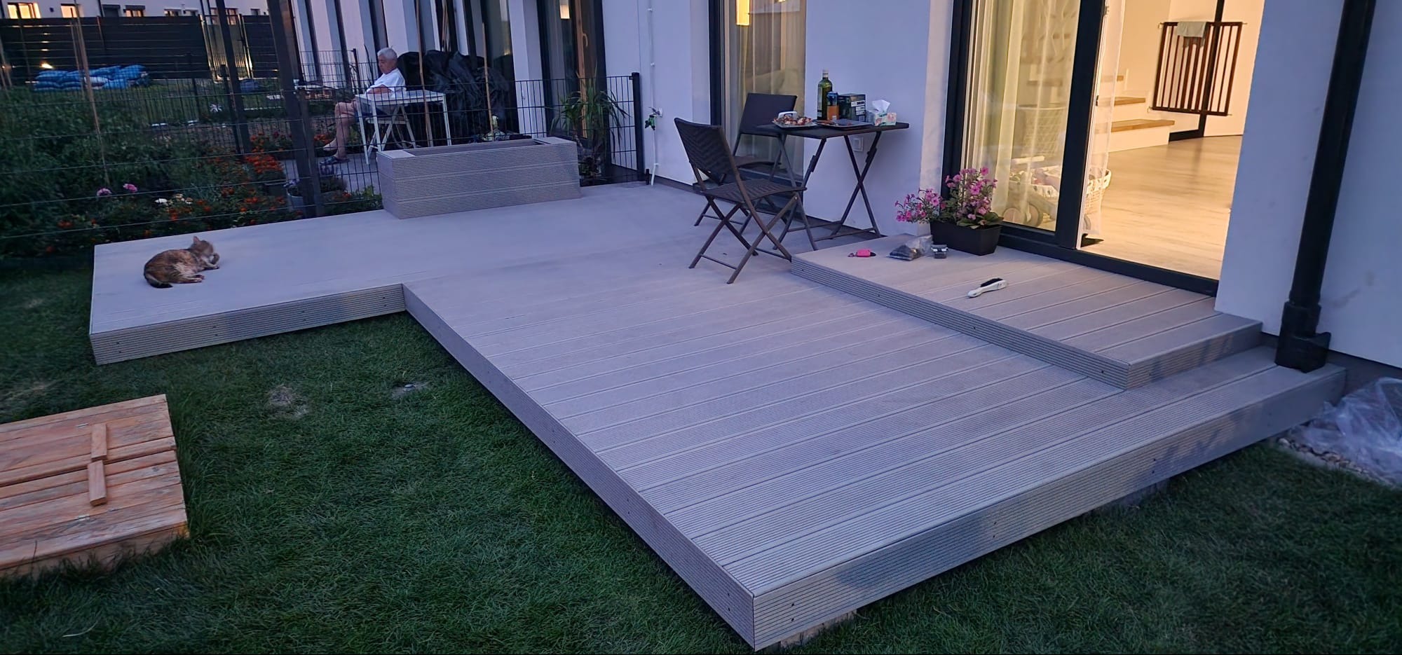 Nowoczesny taras kompozytowy w odcieniach szarości, z niskim stolikiem i krzesłami. Na trawie leży kot. W tle widoczny mężczyzna siedzący przy stole.