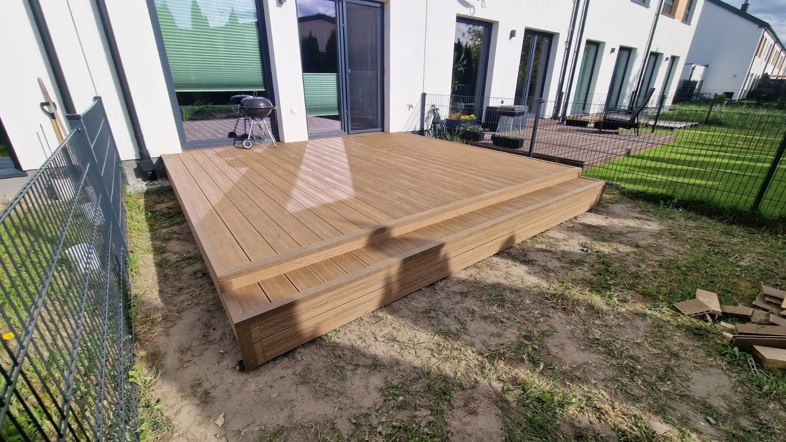 Nowoczesny taras kompozytowy z dwoma stopniami, wykonany z desek w odcieniach brązu, na tle białego budynku z dużymi oknami i zielonego trawnika.