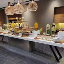 Elegancki stół cateringowy z deserami, ciastami i napojami owocowymi na tle nowoczesnego wnętrza z drewnianymi akcentami i designerskimi lampami.