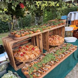 Szulta Catering - Dekoracyjny stół z przekąskami i kwiatami podczas eventu plenerowego. Bruschetty i krokiety na drewnianych deskach i skrzynkach, elegancki catering.