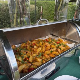 Szulta Catering - Podgrzewacz bufetowy ze smażonymi ziemniakami, posypanymi zieloną pietruszką i czerwoną papryką. W tle dekoracje z kwiatów w wysokich wazonach, zielony trawnik.