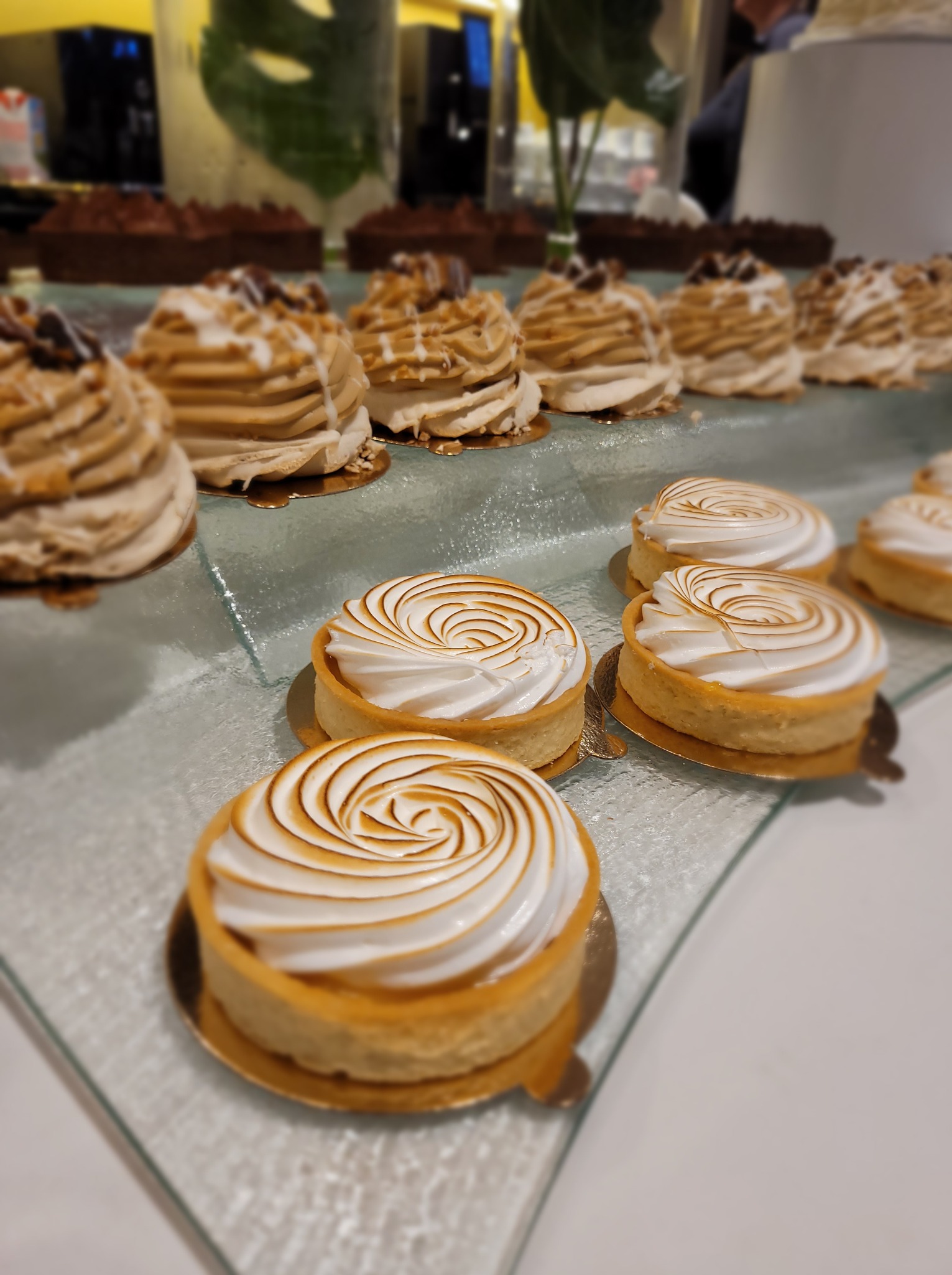 Elegancki bufet deserowy: tarta cytrynowa z bezą i mini torciki na szklanym podeście, w tle roślinna dekoracja. Delikatne oświetlenie podkreśla apetyczny wygląd.