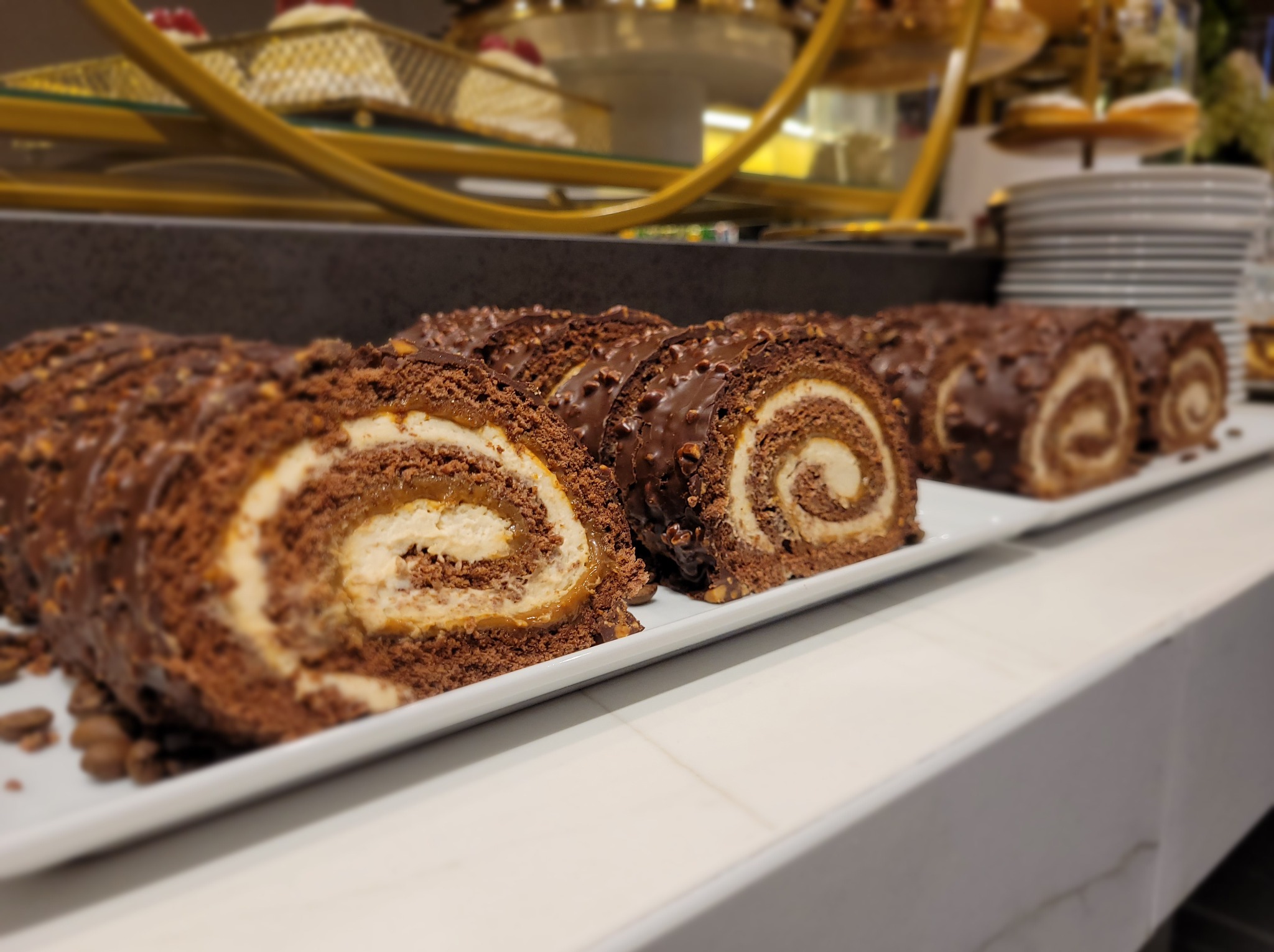 Apetyczne, przekrojone roladki czekoladowe z kremowym nadzieniem na białym talerzu. W tle widoczne inne desery na paterach, sugerujące słodki stół.