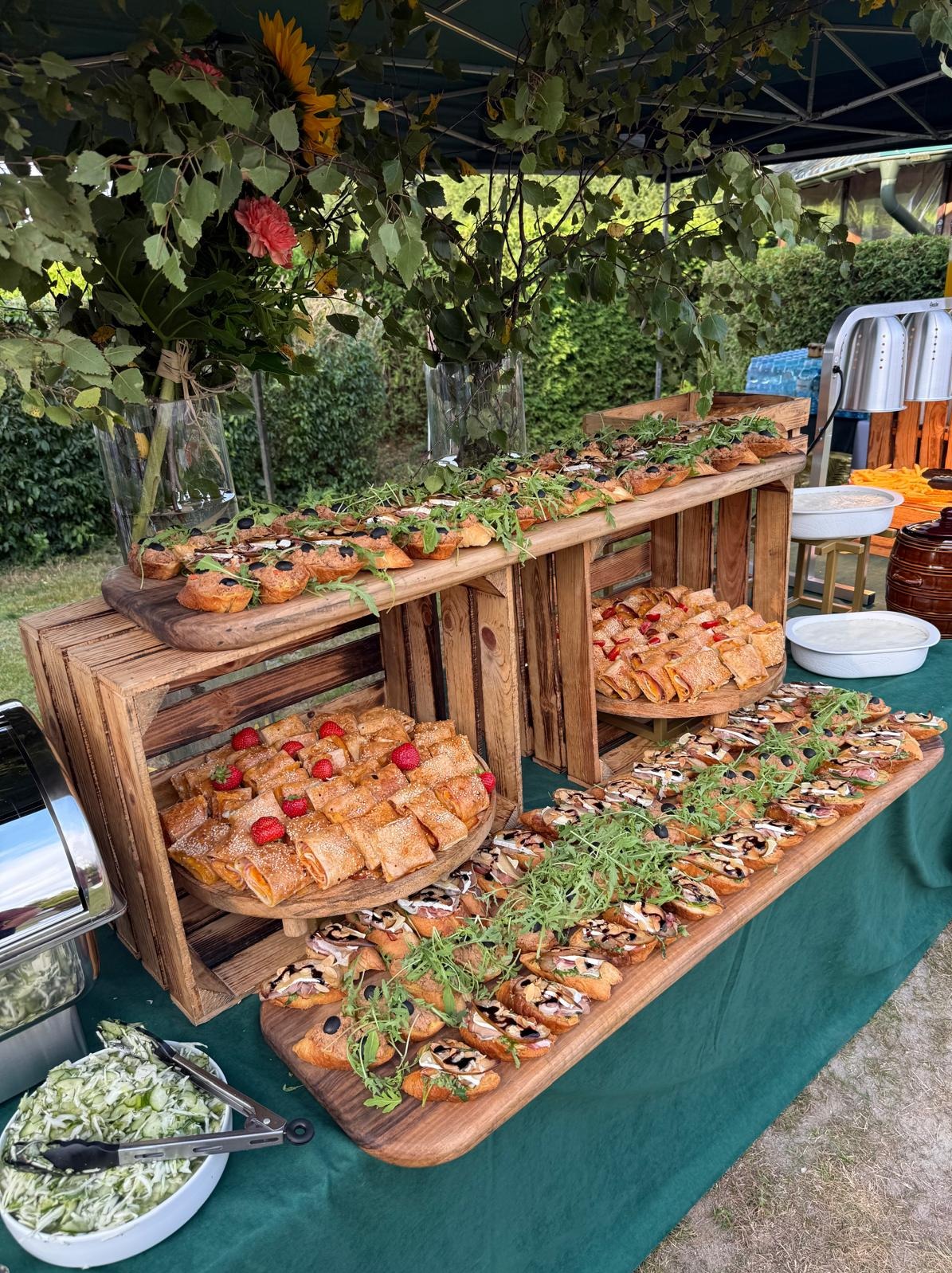 Dekoracyjny stół z przekąskami i kwiatami podczas eventu plenerowego. Bruschetty i krokiety na drewnianych deskach i skrzynkach, elegancki catering.