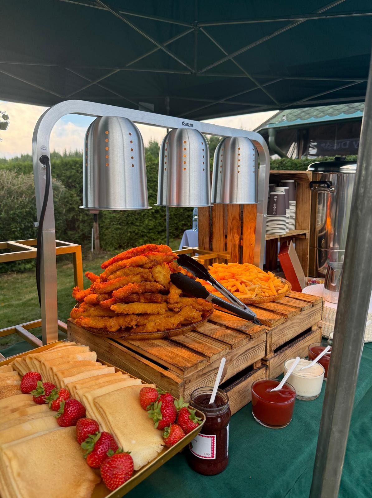 Bufet z podgrzewaczami: naleśniki z truskawkami, frytki, nuggetsy. Drewniane skrzynki, sosy i dżem. Catering plenerowy pod namiotem.