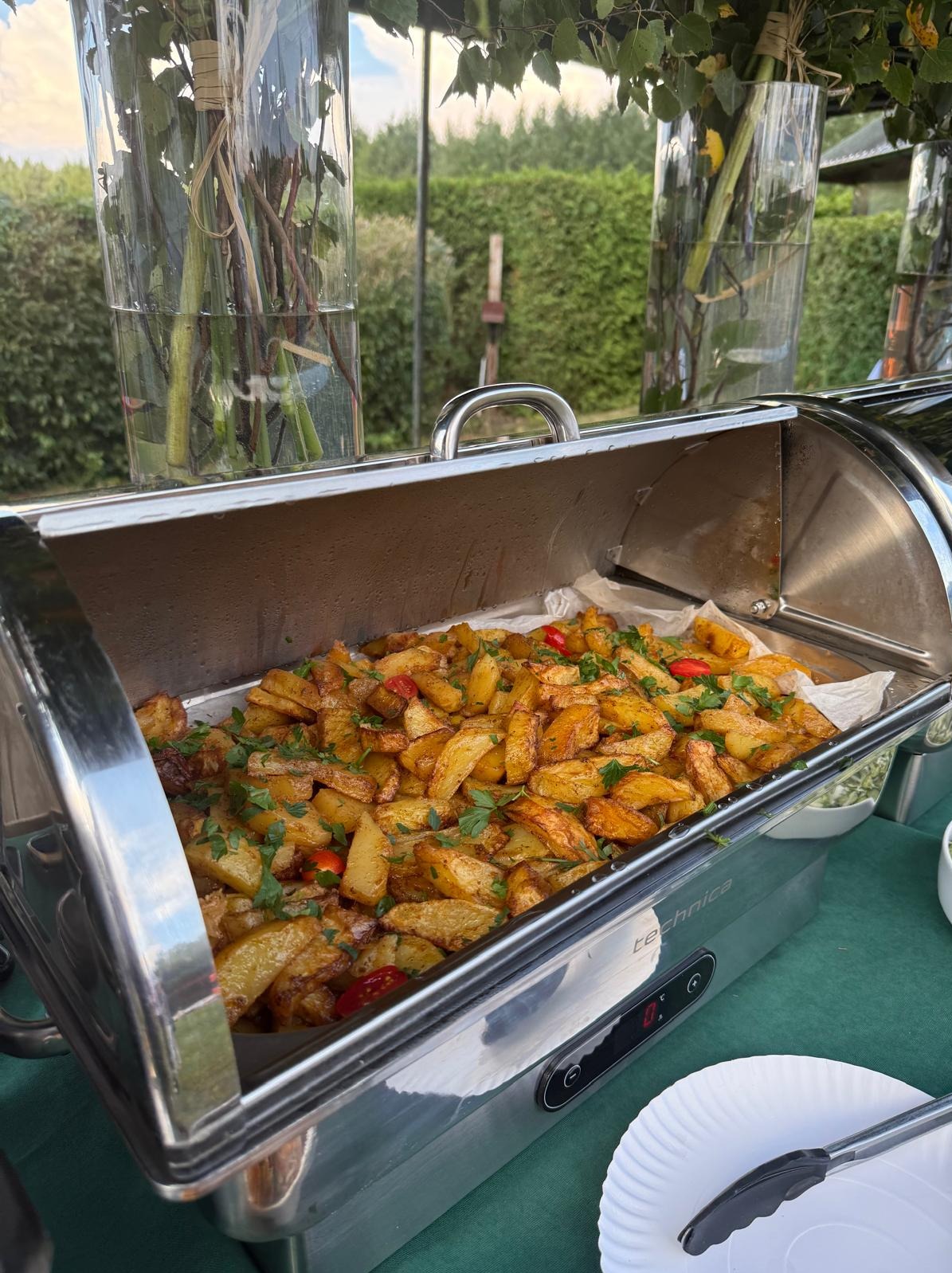 Podgrzewacz bufetowy ze smażonymi ziemniakami, posypanymi zieloną pietruszką i czerwoną papryką. W tle dekoracje z kwiatów w wysokich wazonach, zielony trawnik.