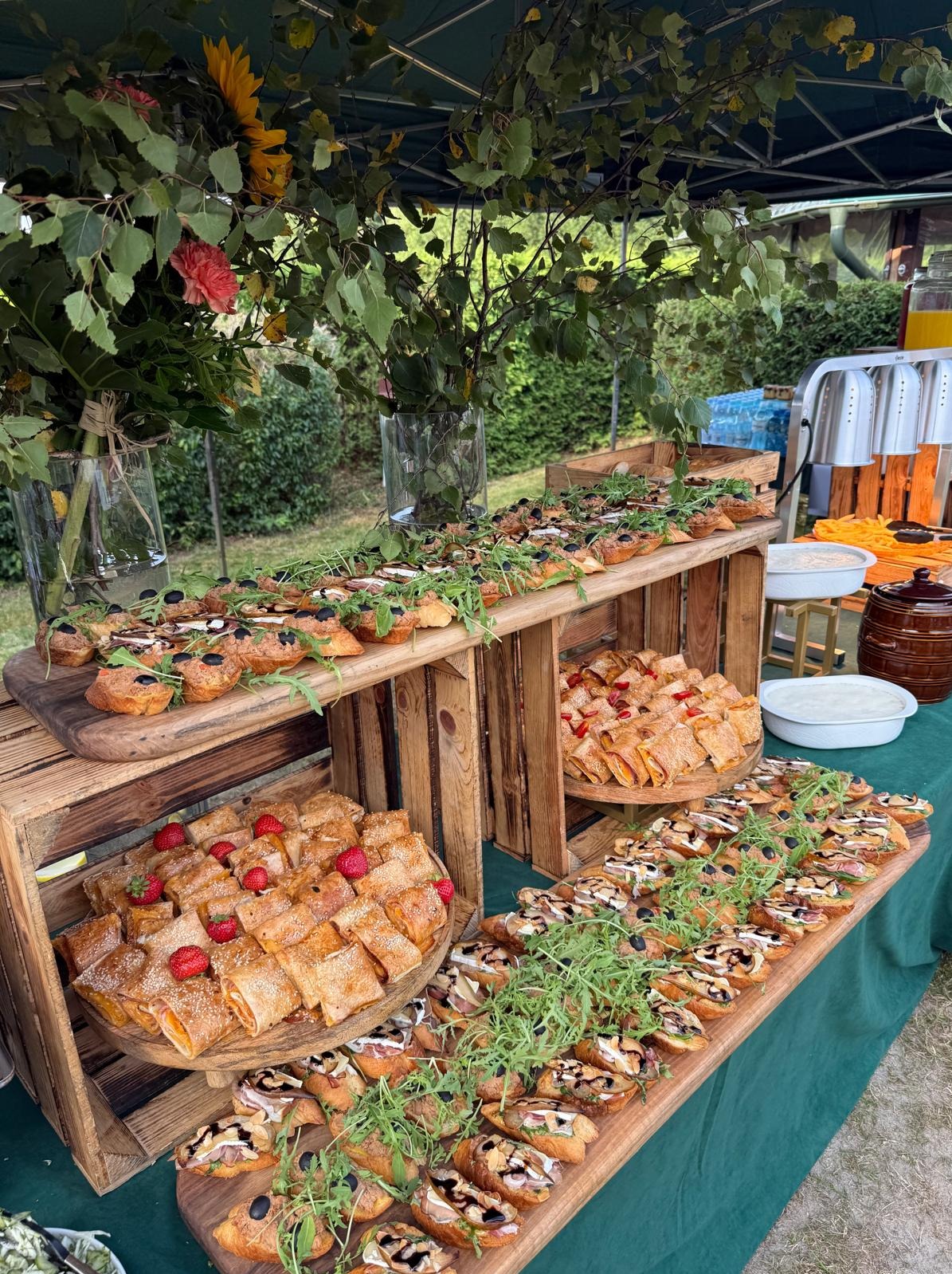 Stół z przekąskami na przyjęciu plenerowym: bruschetty z rukolą i oliwkami, roladki z truskawkami. Dekoracja z kwiatów i zieleni. Bufet z napojami w tle.