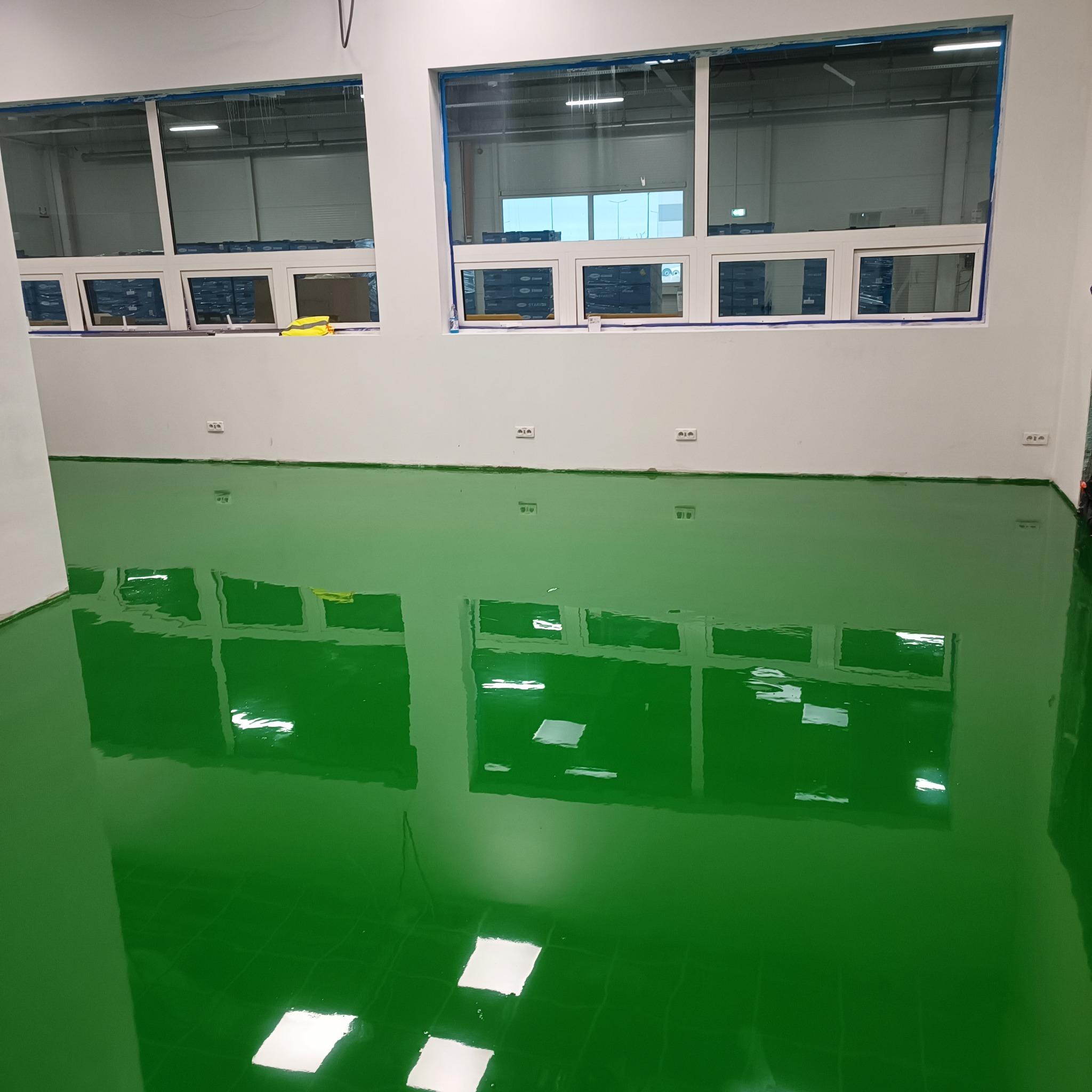 Błyszcząca, zielona posadzka żywiczna w hali produkcyjnej, z odbiciem okien. Widoczne taśmy malarskie zabezpieczające okna. Gładka powierzchnia.