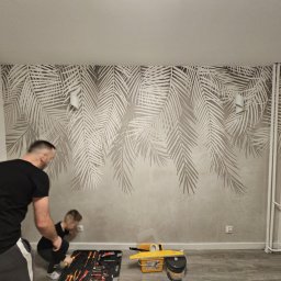 ARKADIUSZ Łapiński - Tapetowanie ściany z motywem liści palmowych. Mężczyzna i dziecko pracują przy gniazdku. Widoczne narzędzia: skrzynka z narzędziami, wiadro i poziomica.