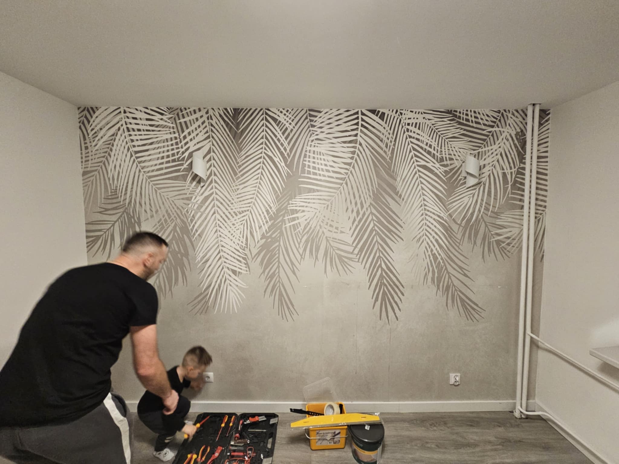 Tapetowanie ściany z motywem liści palmowych. Mężczyzna i dziecko pracują przy gniazdku. Widoczne narzędzia: skrzynka z narzędziami, wiadro i poziomica.