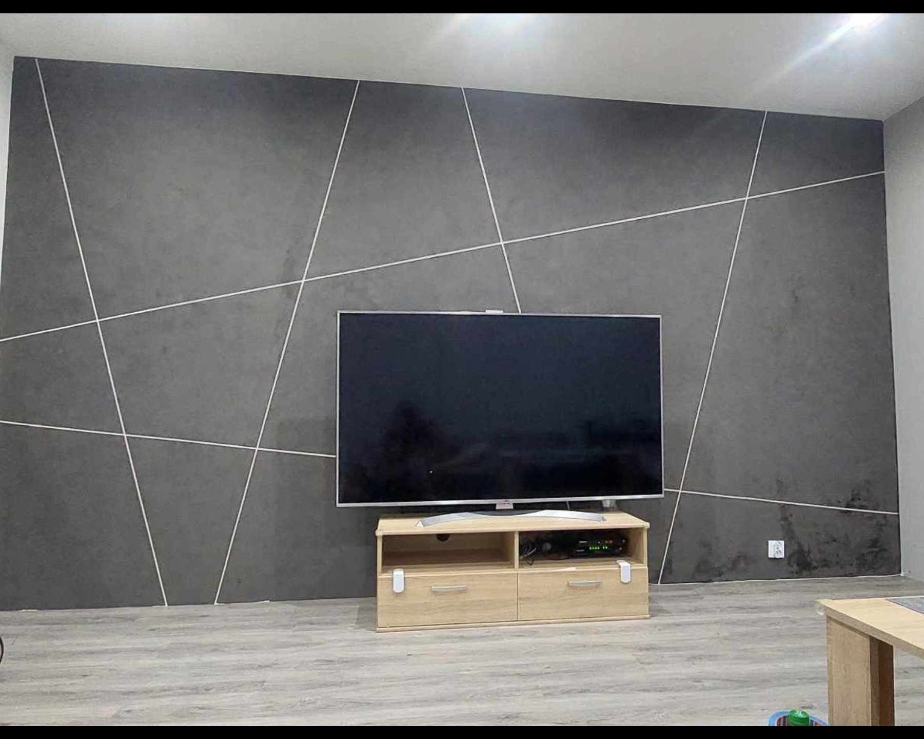 Szara ściana z geometrycznym wzorem z białych linii, podświetlona telewizorem na drewnianej szafce RTV. Nowoczesne wykończenie wnętrza salonu.