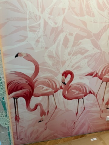 Różowa tapeta z motywem flamingów i liści, pokrywająca ścianę w pomieszczeniu. Widoczne detale wzoru i faktura tapety. Dekoracja wnętrza w pastelowych barwach.
