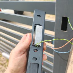 Tomasz Piecyk voltom instalacje elektryczne pomiary - Montaż elektrozaczepu w metalowej bramie posesji. Widoczne przewody i element zamka w dłoni montera, gotowy do instalacji w słupku bramy.