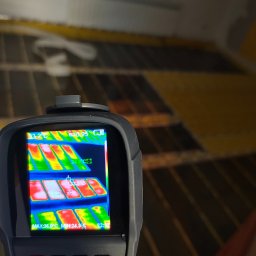 Tomasz Piecyk voltom instalacje elektryczne pomiary - Termowizja ogrzewania podłogowego w trakcie testów. Na ekranie urządzenia widoczne rozłożenie temperatur. Żółte rury na podłożu w tle.